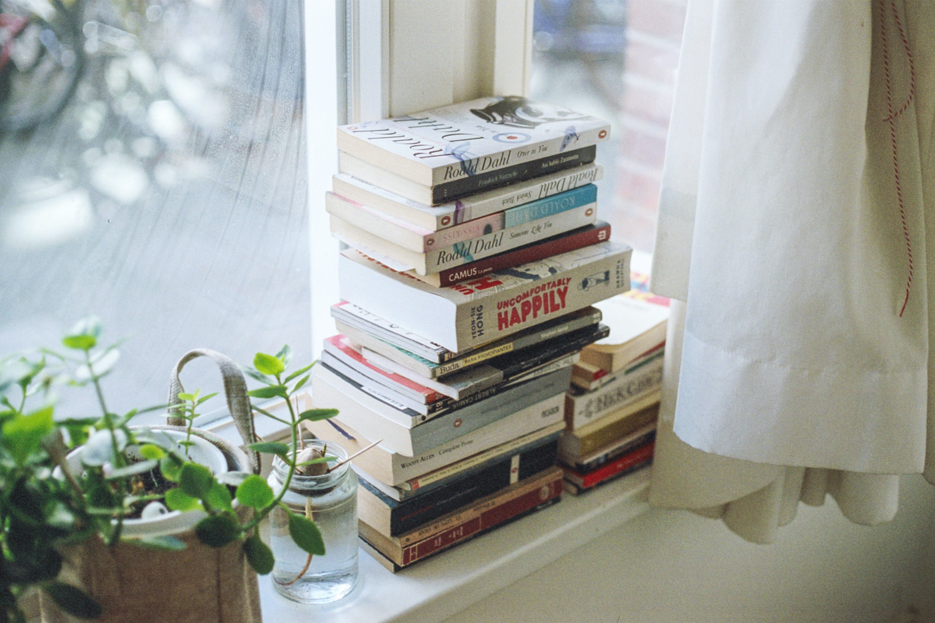 A stack of various books on top of a windowsill. Beside the stack of books are a few houseplants.